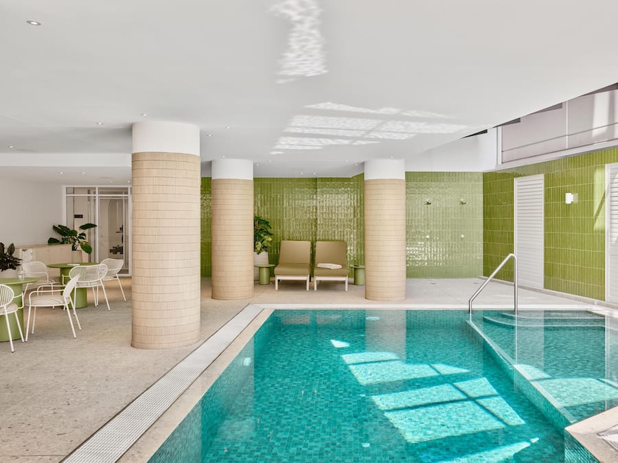 A modern indoor pool area with a clear pool, beige pillars, green tiled walls, and a seating area with a sofa and chairs.