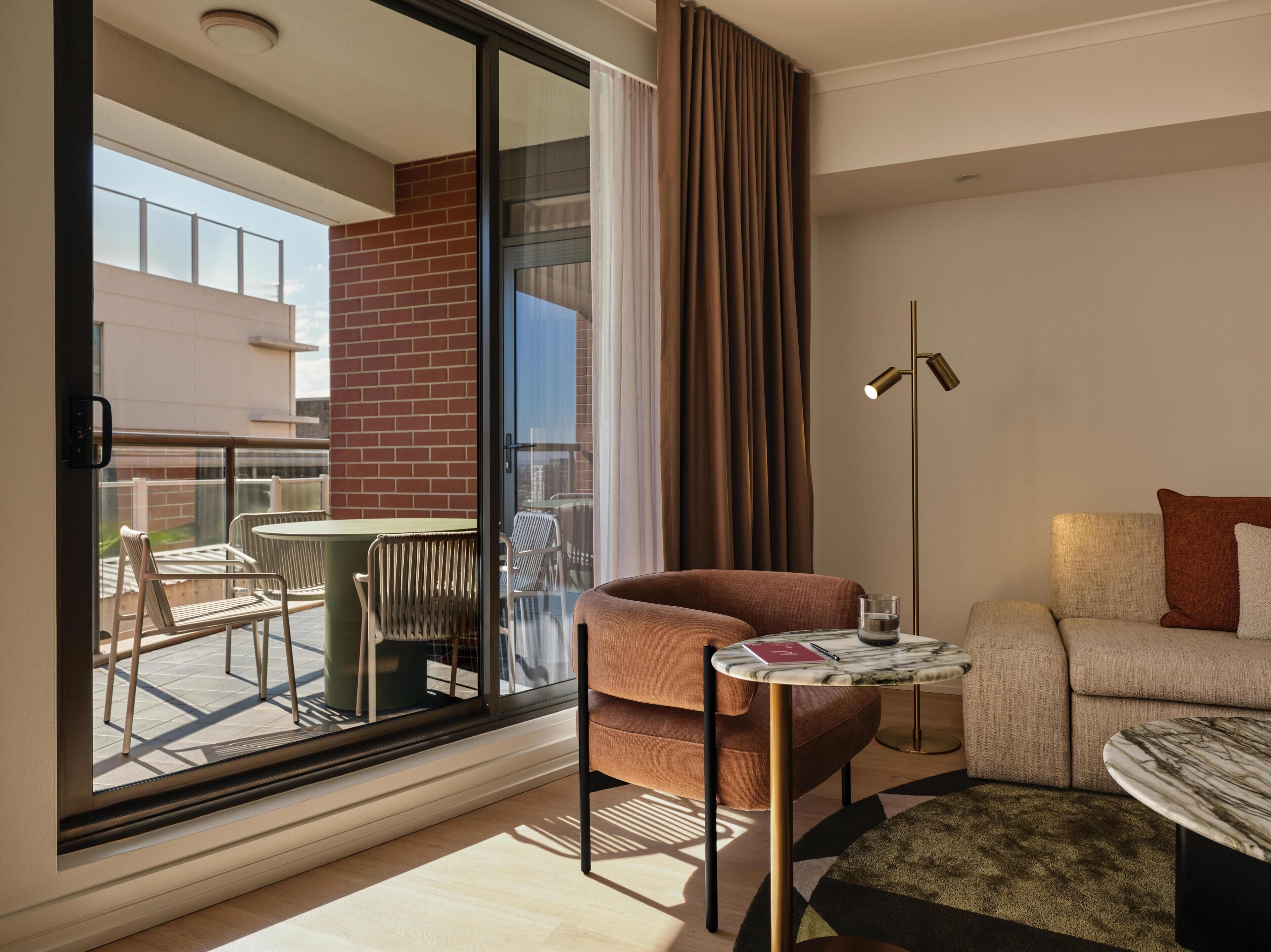 Una sala moderna con un sofá, una mesita y una silla. Se ve un balcón con mobiliario exterior a través de una puerta de cristal.