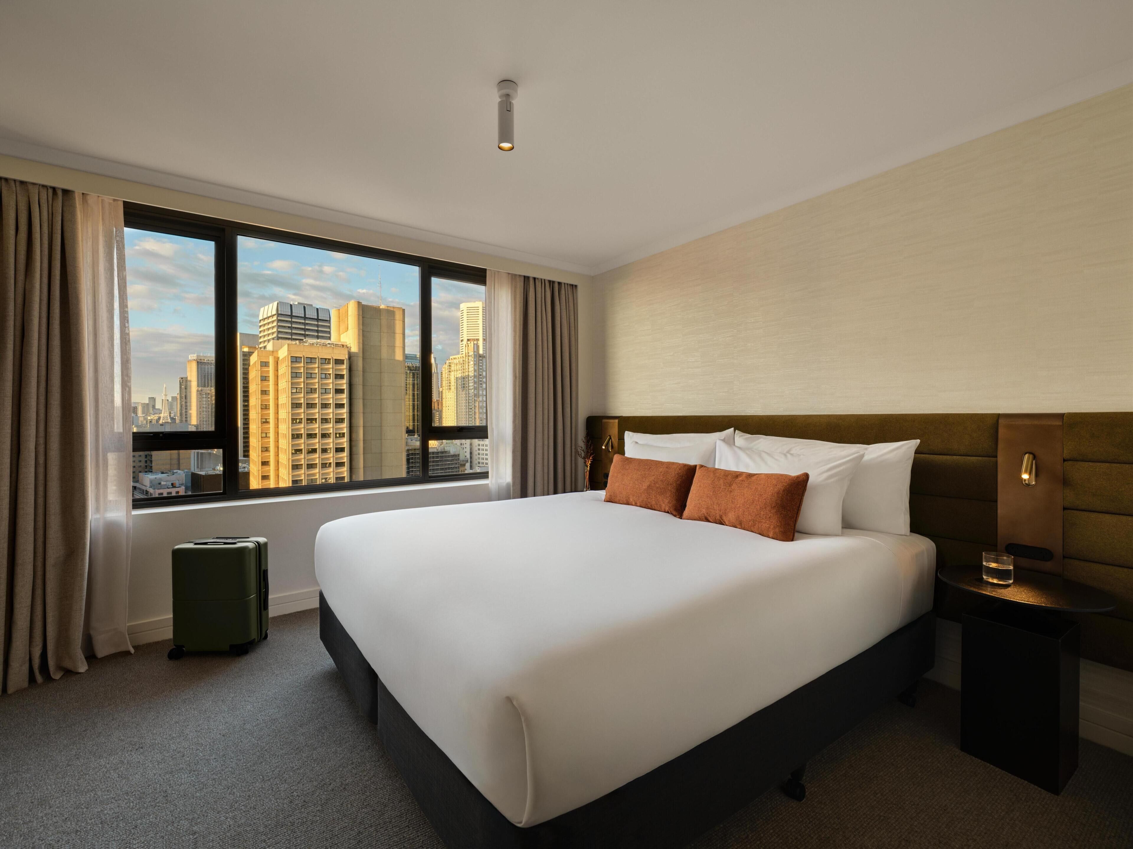 A hotel room with a large bed, a nightstand, a lamp, a suitcase, and a view of a cityscape through a large window.