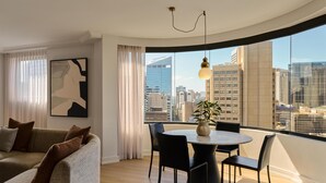 A modern living room with a sofa, a dining table with chairs, a large window with a city view, and a pendant light.