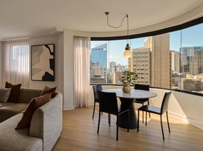 A modern living room with a sofa, a dining table with chairs, a large window with a city view, and a pendant light. - Adina Sydney Town Hall (Sydney)
