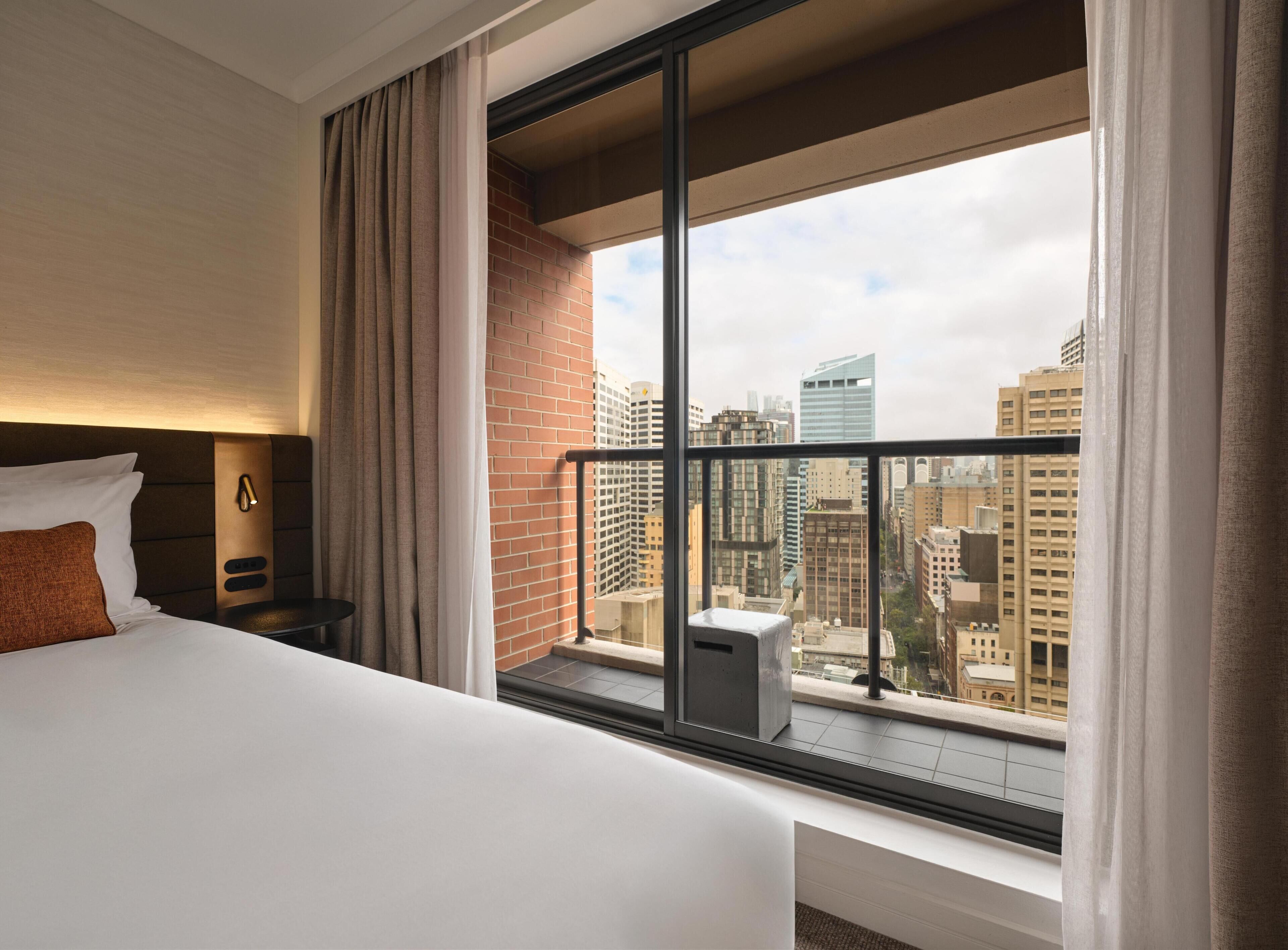 Habitación de hotel con un ventanal grande, una cama, una mesita de noche y vistas a edificios de la ciudad.