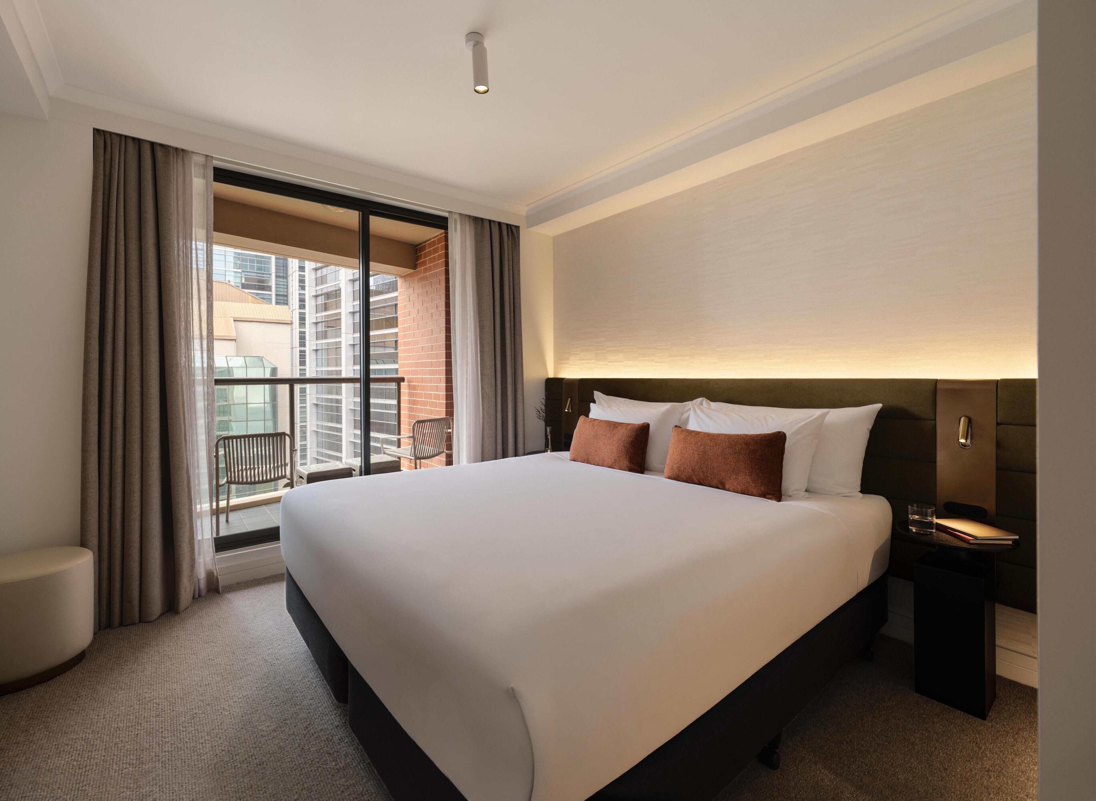 A hotel room with a large bed, a nightstand, a chair, and a view of a cityscape through a balcony.