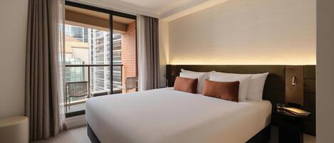 A hotel room with a large bed, a nightstand, a chair, and a view of a cityscape through a balcony.