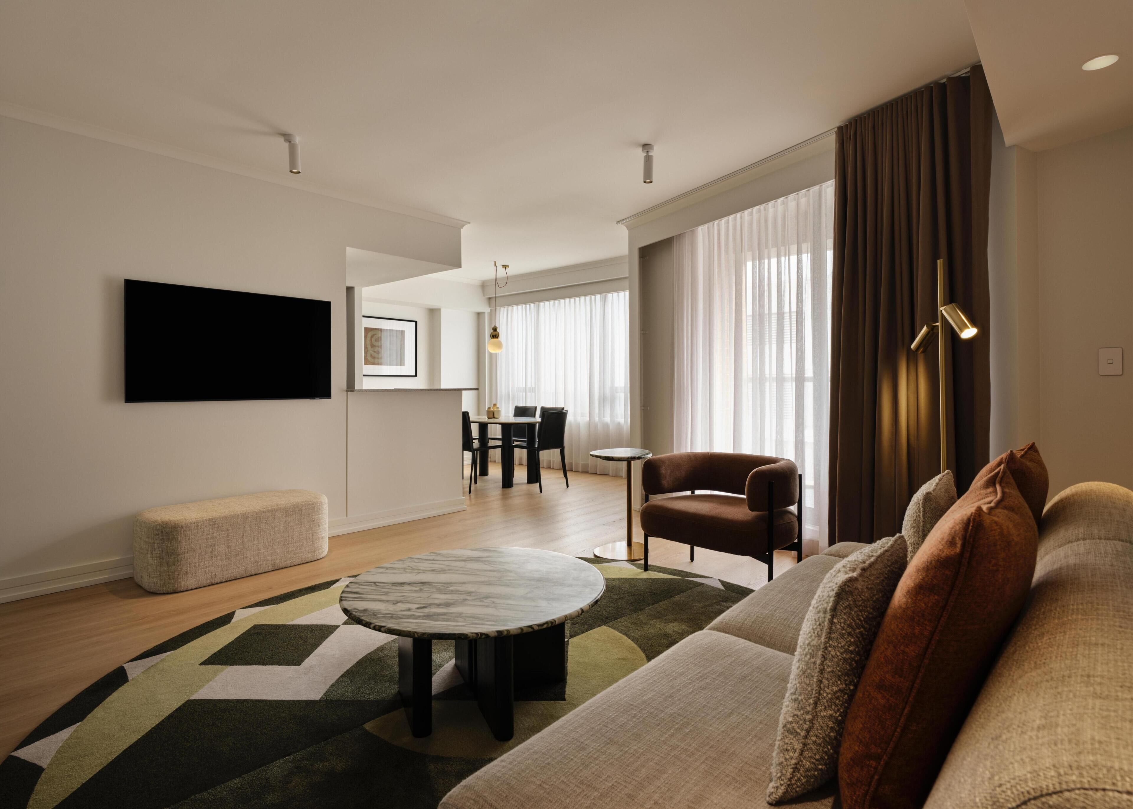 Una sala de estar moderna con un sofá, una mesa de centro, un televisor montado en la pared y un comedor al fondo.