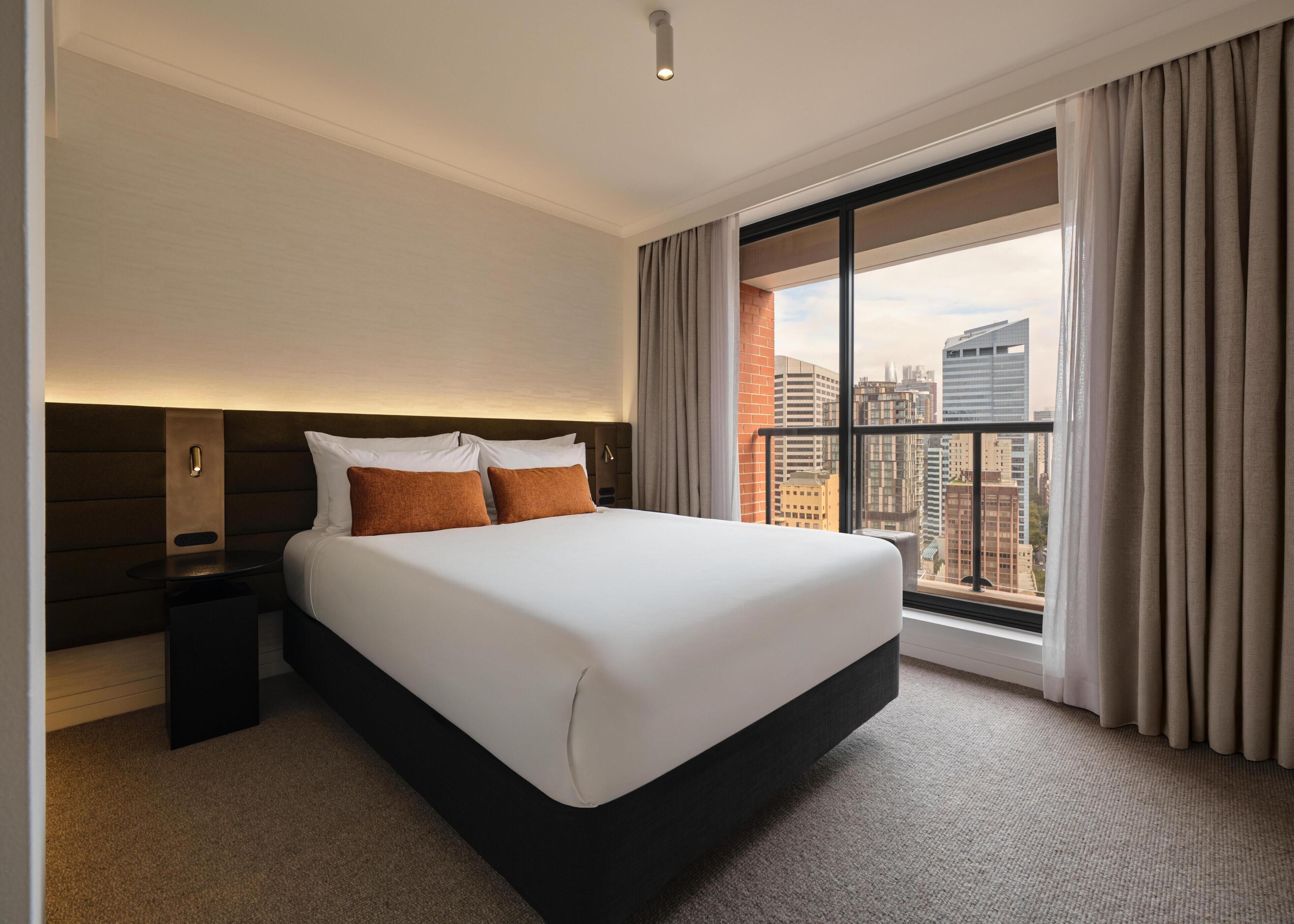 Habitación de hotel con una cama grande, una mesita de noche, una lámpara de pared y vistas de la ciudad a través de un ventanal.