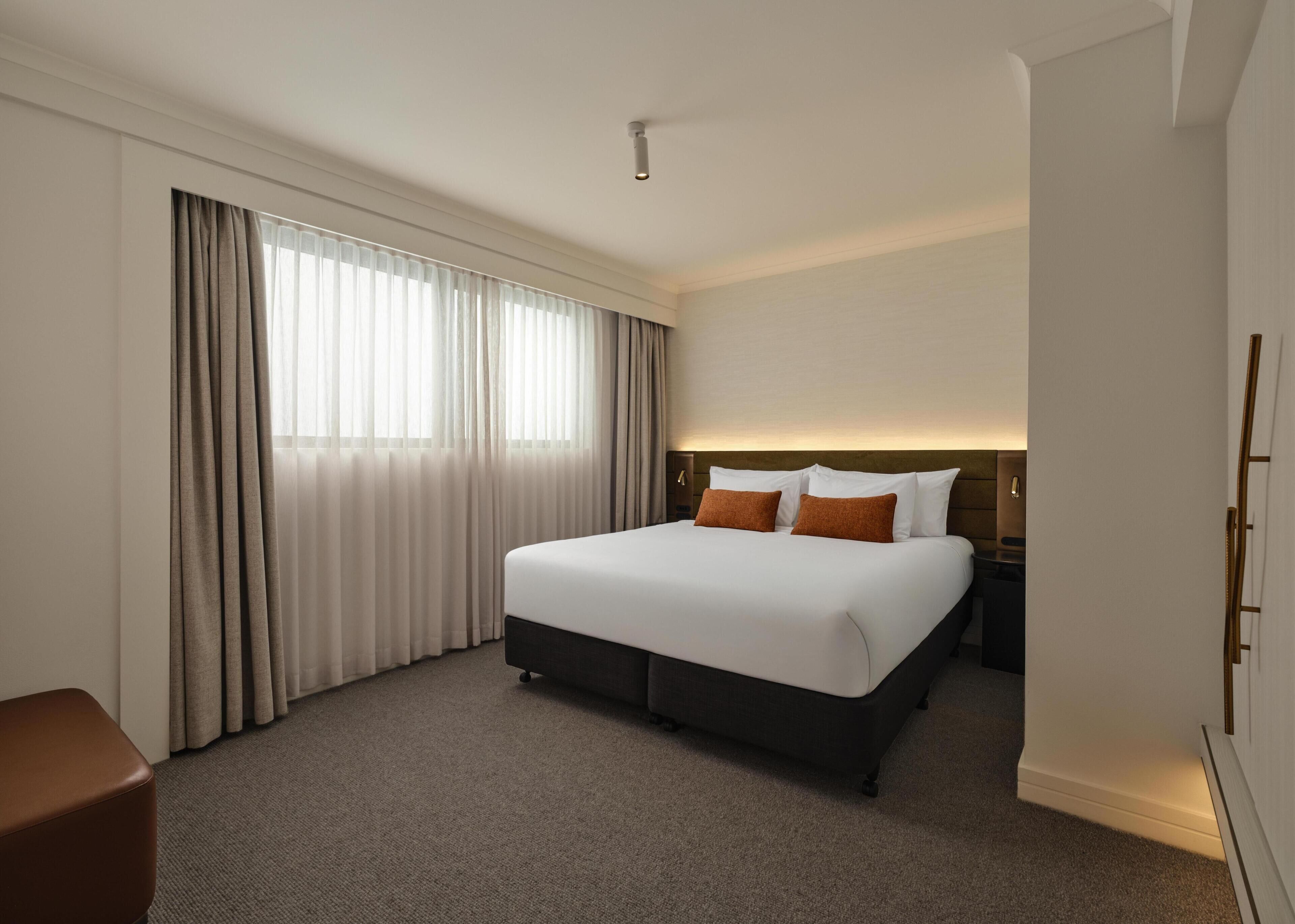 Habitación de hotel con una cama grande, una mesita de noche, una lámpara, una ventana con cortinas y una puerta.