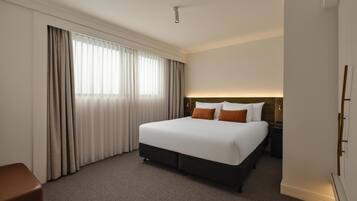 Habitación de hotel con una cama grande, una mesita de noche, una lámpara, una ventana con cortinas y una puerta.