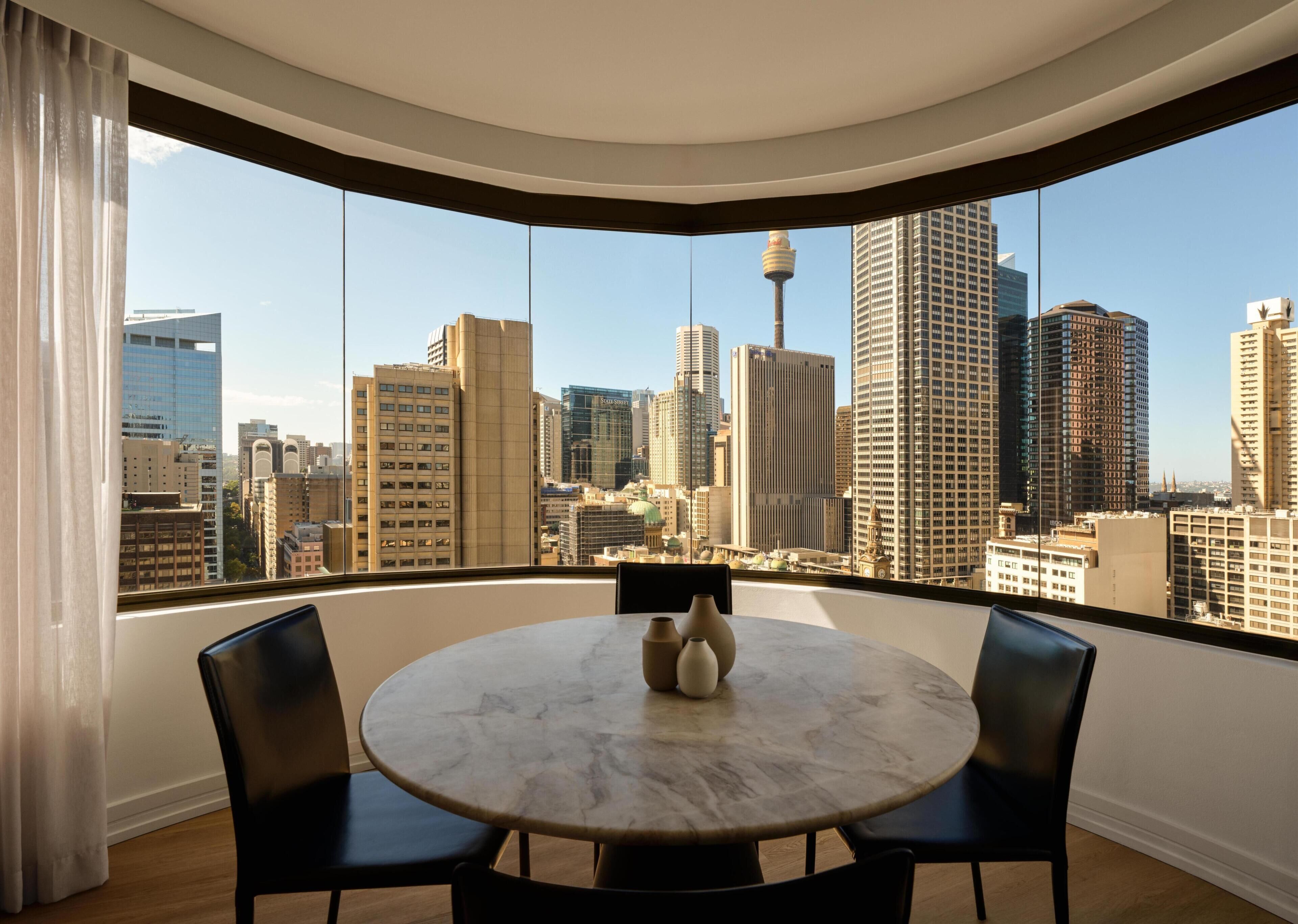 A round table with chairs, a vase, and a cityscape view through large windows.