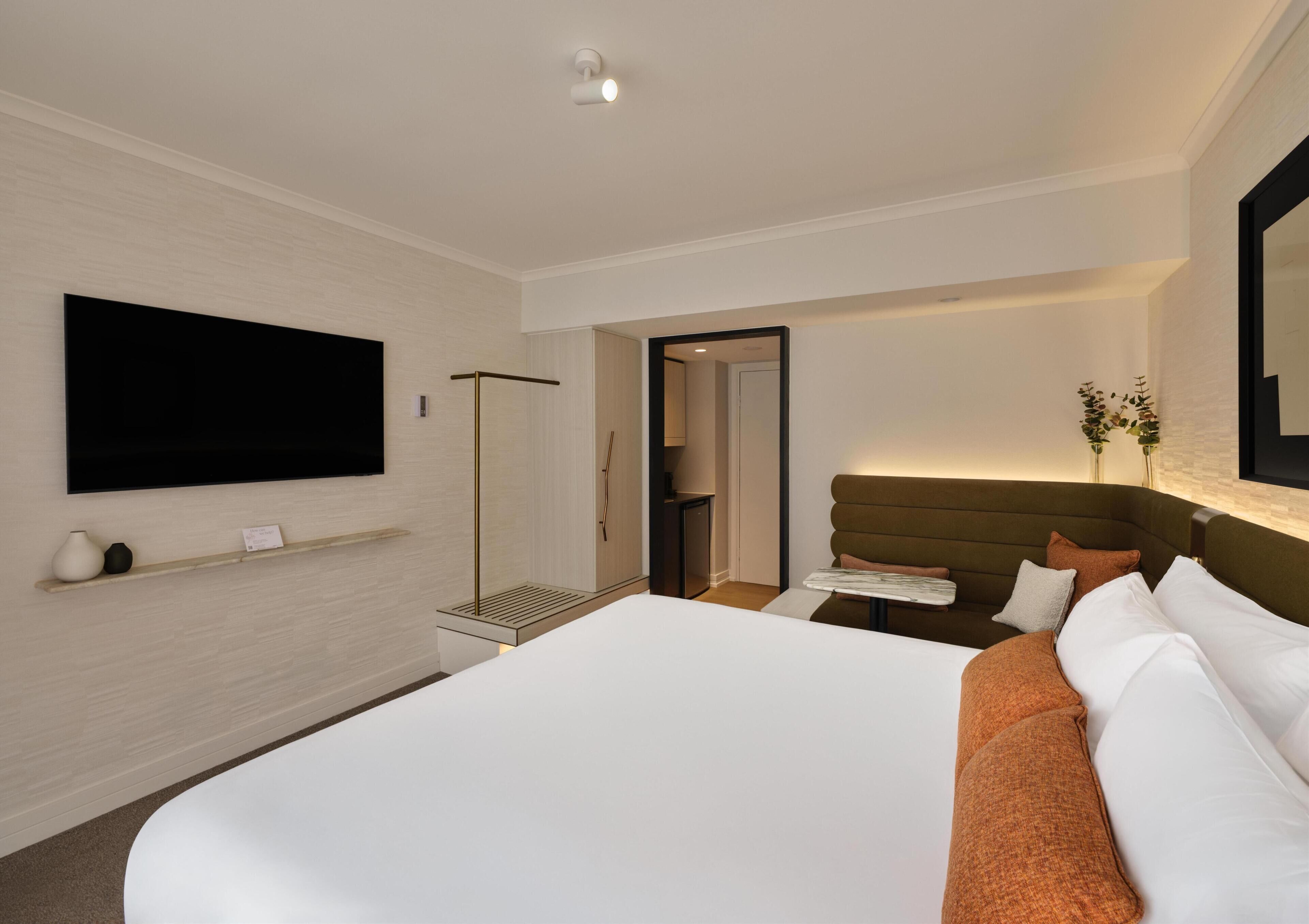 Quarto de hotel com uma cama grande, televisão de tela plana na parede, mesa de cabeceira e porta de acesso a um banheiro.