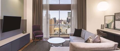 A modern living room with a large TV, comfortable seating, a small table, and a city view through large windows.