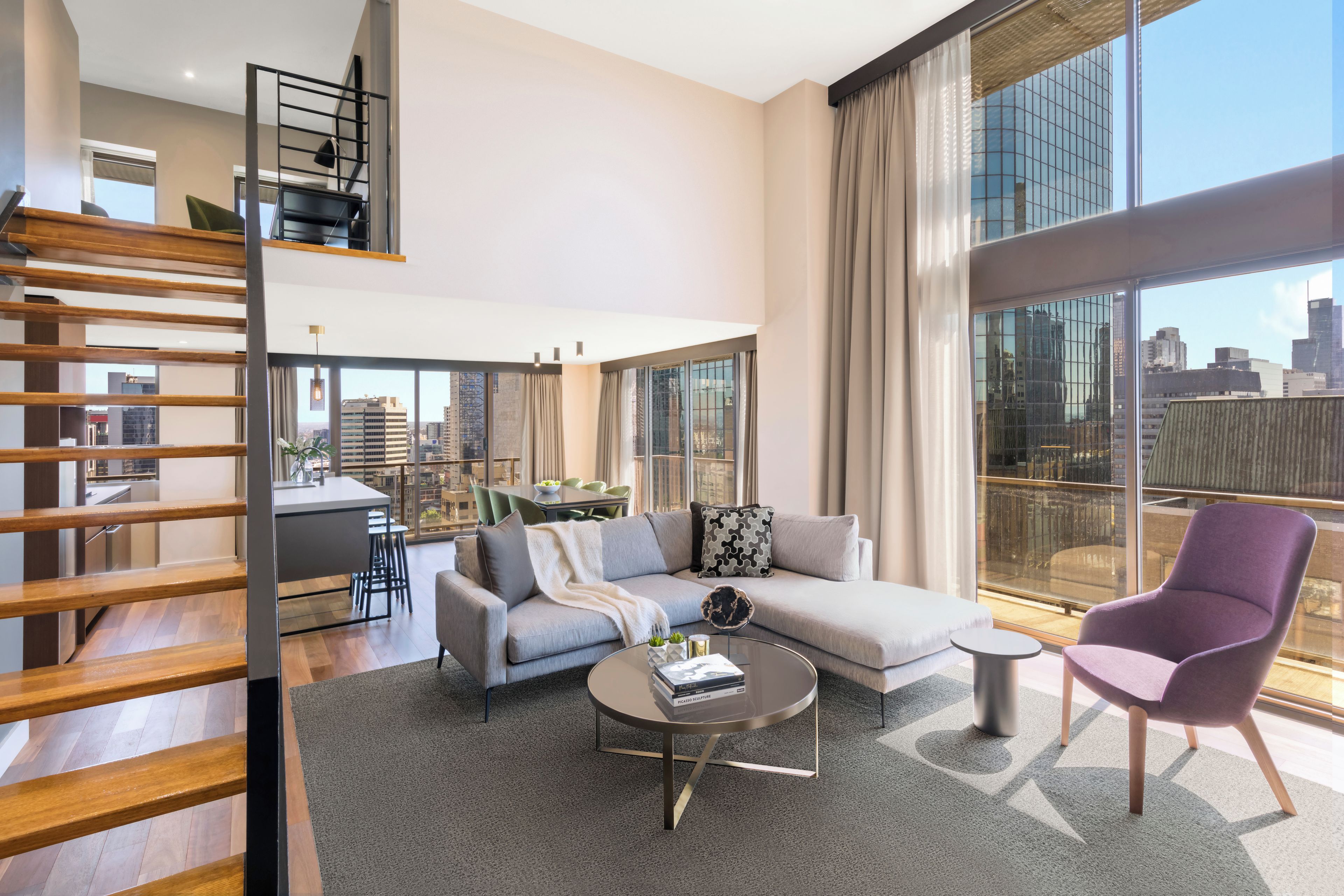 Una sala moderna con un sofá, una mesa de centro y un sillón morado. Se ve una escalera a la izquierda y vistas de la ciudad a través de amplios ventanales.
