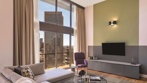Un salon moderne avec un canapé, un fauteuil et une table basse, un téléviseur à écran plat et une vue sur la ville grâce à de grandes fenêtres.