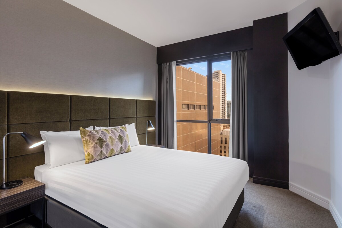 Habitación de hotel con una cama grande, una mesita de noche, un escritorio, un televisor de pantalla plana en la pared y una ventana con cortinas.