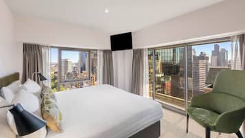 A hotel room with a large bed, a green armchair, and a city view through floor-to-ceiling windows.