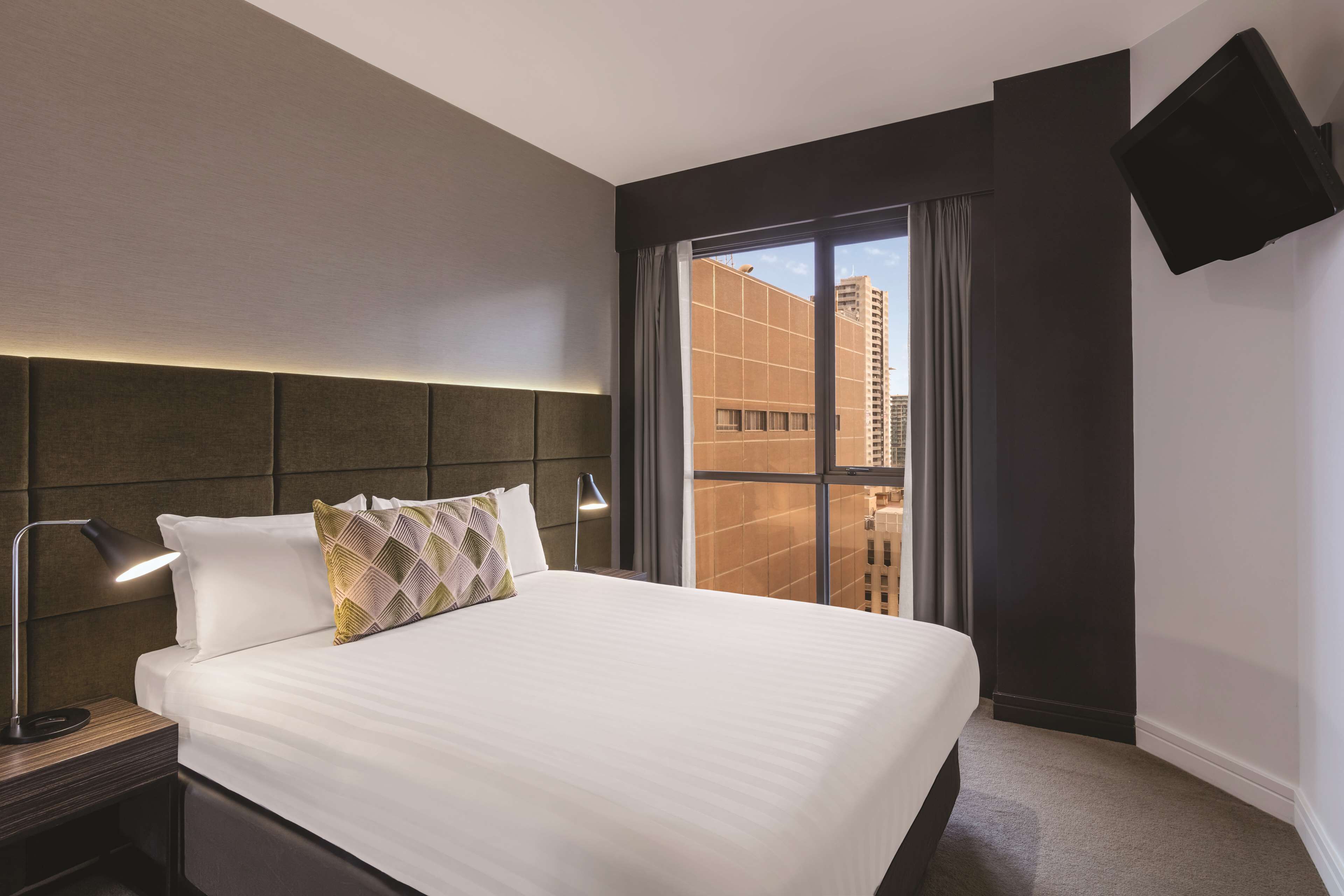 Ein Hotelzimmer mit einem großen Bett, einem Kopfteil, einem Nachttisch mit Lampe, einem an der Wand befestigten Flachbildfernseher und Blick auf Gebäude durch ein Fenster.