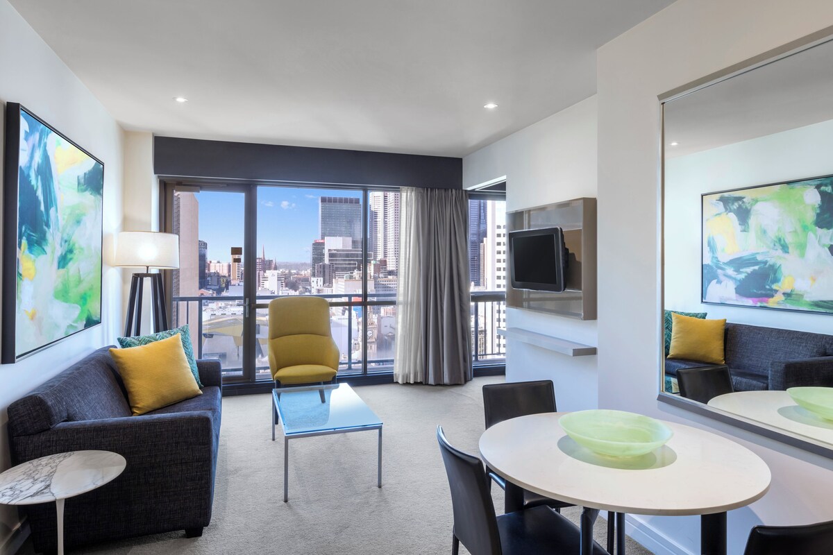 Habitación de hotel moderna con vistas a la ciudad, un sofá, una silla, una mesa de comedor y un televisor montado en la pared.