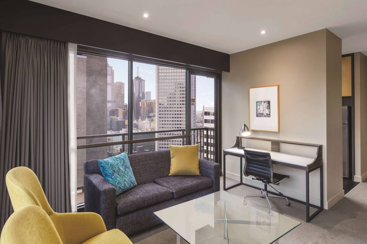 A modern hotel room with a city view, a sofa, a desk, and a glass coffee table.