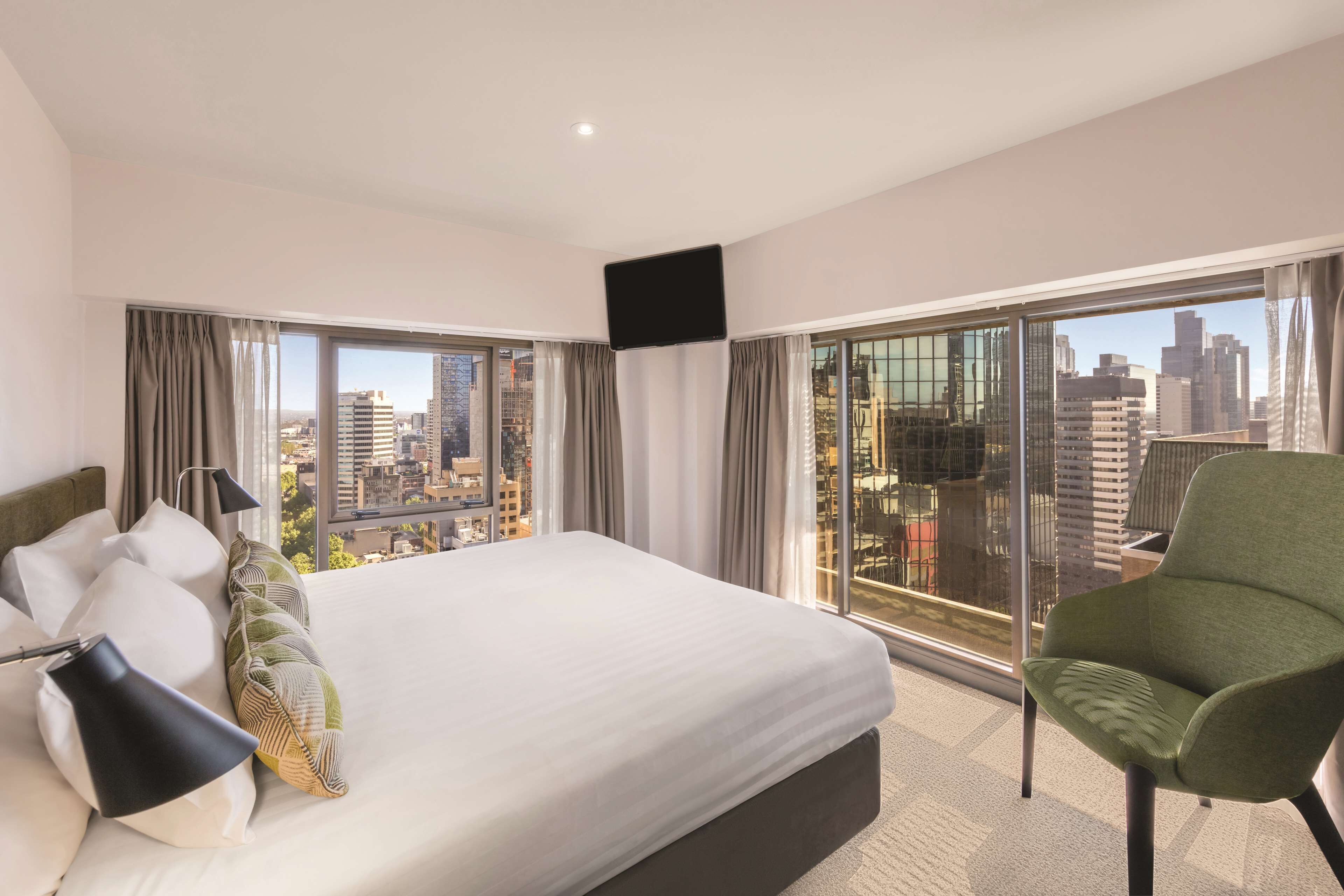 Una habitación de hotel con una cama grande, una silla verde y vistas a la ciudad a través de amplias ventanas.