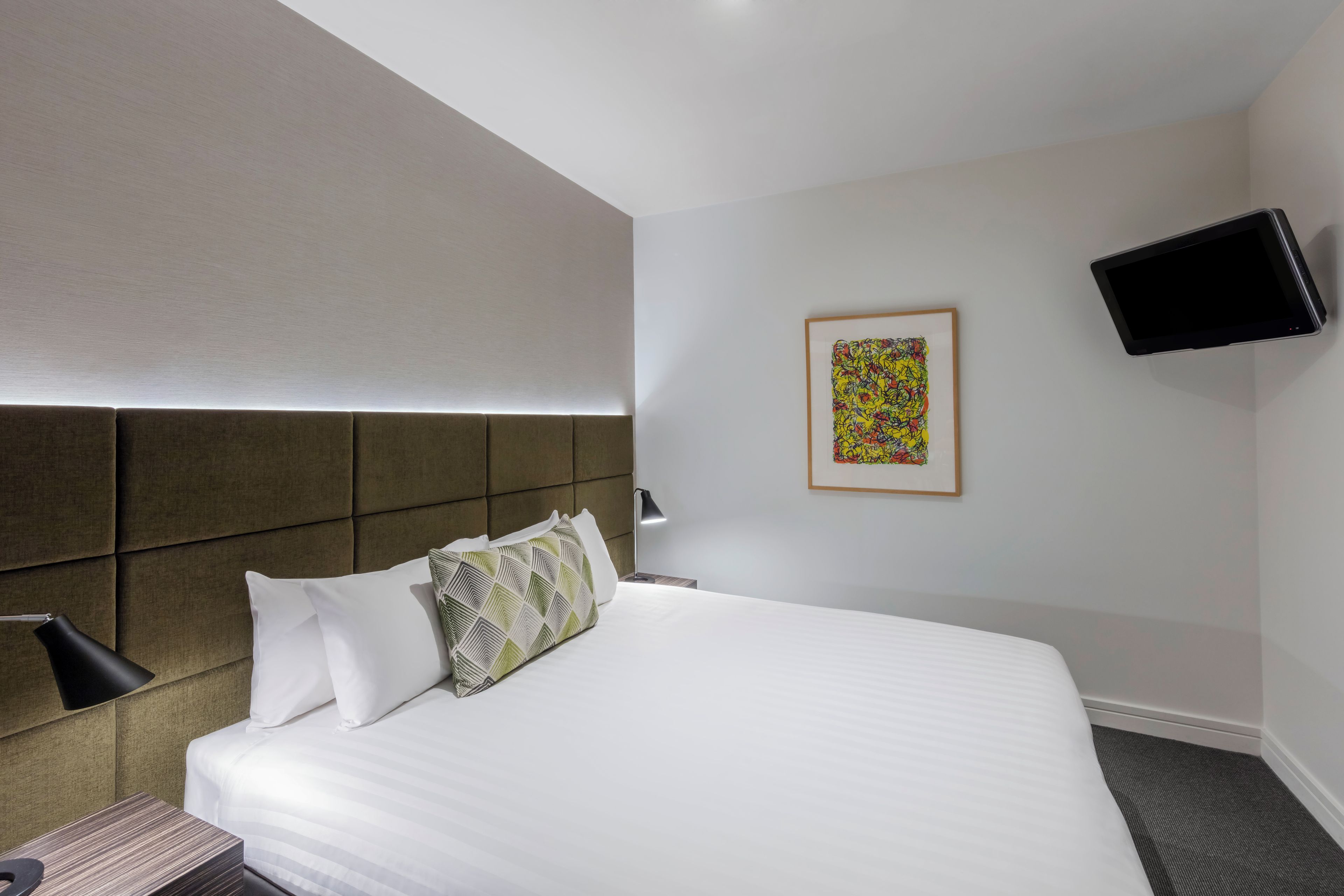 A hotel room with a bed, a headboard, a bedside table, a lamp, a flat-screen TV mounted on the wall, and a framed artwork.