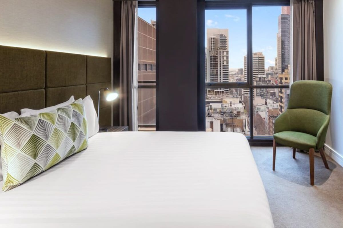 Hotelkamer met een groot bed, een groene fauteuil, een flatscreen televisie aan de wand en uitzicht op de stad door grote ramen.