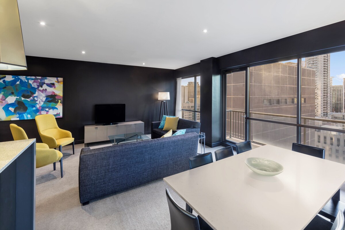 Una habitación de hotel moderna con un ventanal grande, un televisor de pantalla plana, un sofá, una mesa de comedor y un cuadro abstracto colorido en la pared.