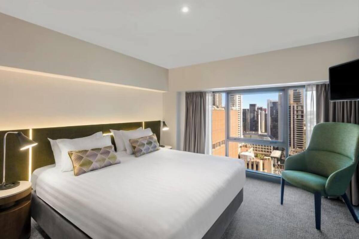A hotel room with a large bed, a green armchair, a TV, and a city view.