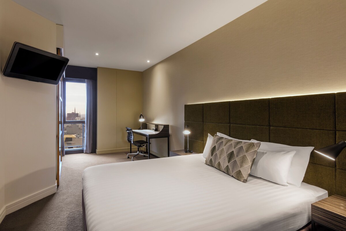 Habitación de hotel con una cama grande, un escritorio, una silla, un televisor de pantalla plana montado en la pared y vistas de la ciudad a través de la ventana.
