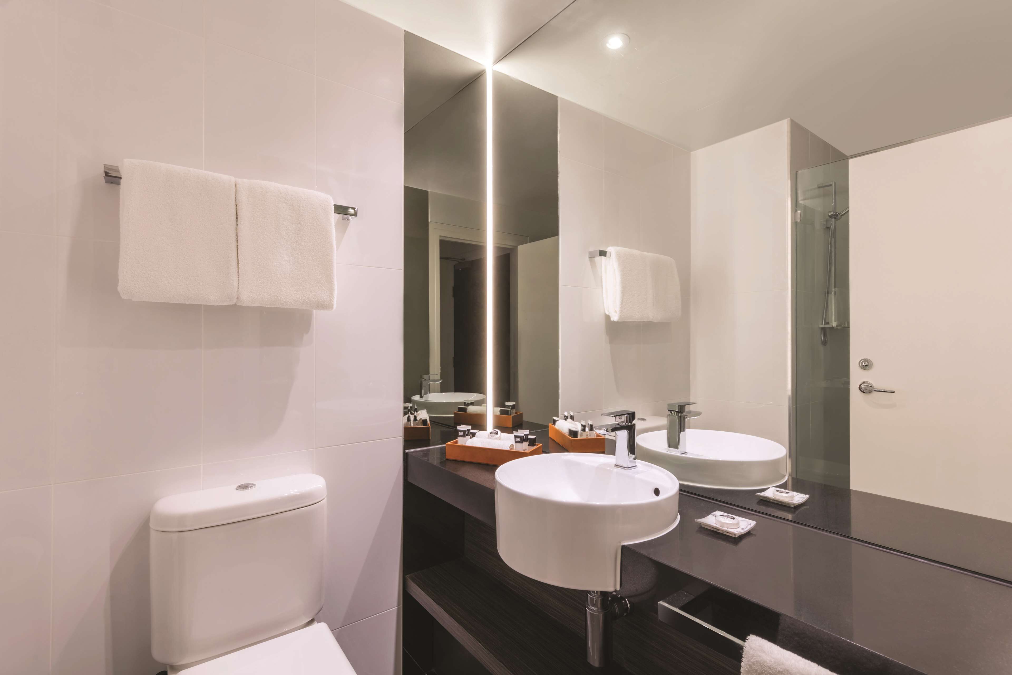 Un baño moderno con inodoro, dos lavamanos, un espejo grande y cabina de ducha de vidrio.