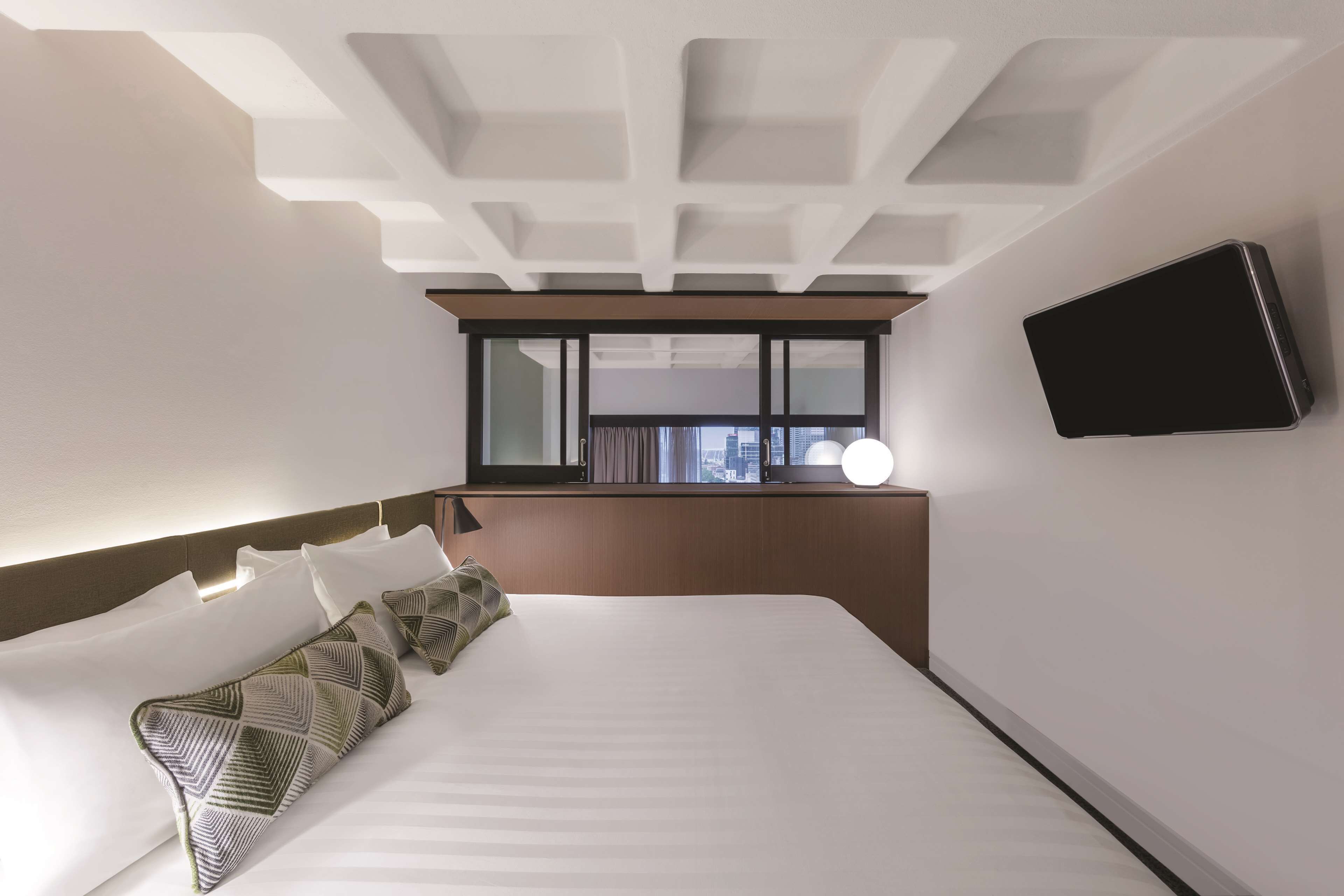 A modern hotel room with a large bed, a flat-screen TV mounted on the wall, and a window with a city view.