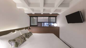 A modern hotel room with a large bed, a flat-screen TV mounted on the wall, and a window with a city view.