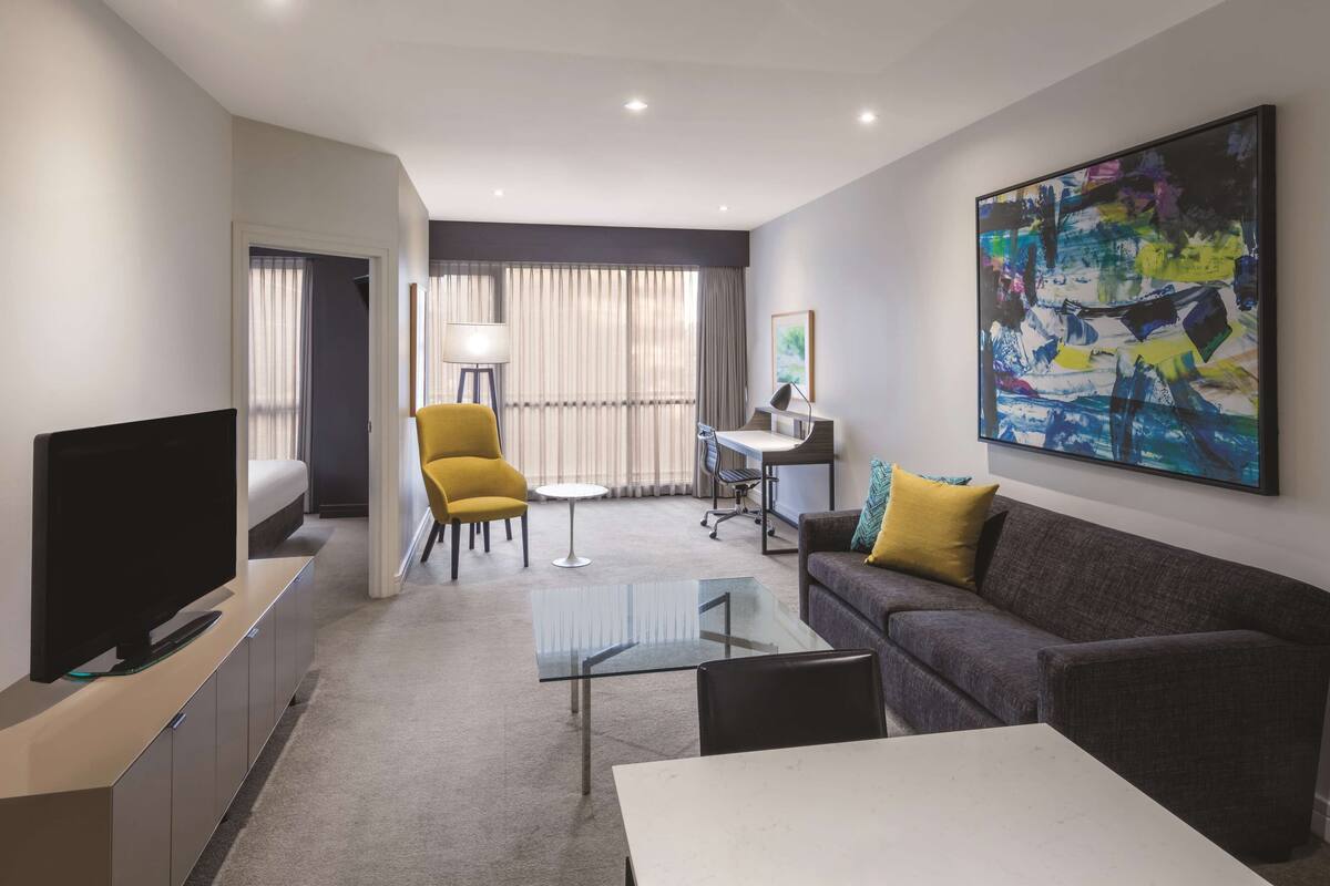 A modern hotel room with a flat-screen TV, a sofa with yellow cushions, a glass coffee table, a small desk with a lamp, and a large abstract painting on the wall.