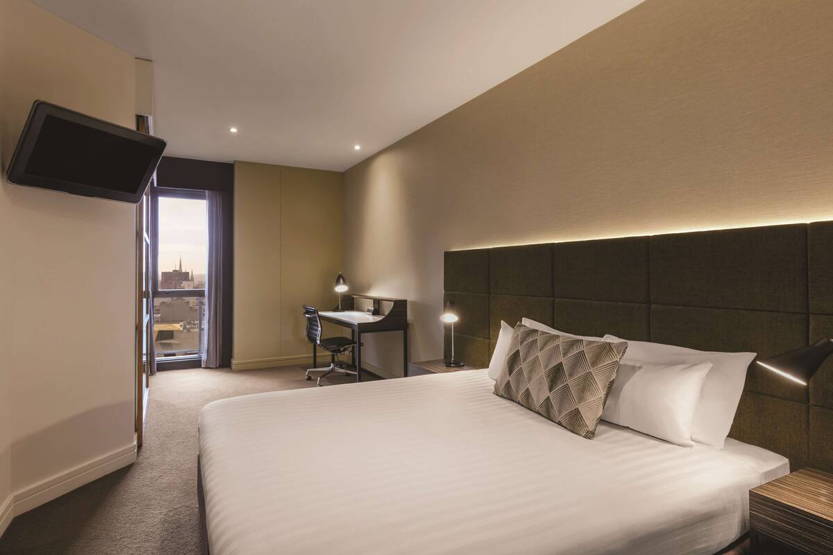 A hotel room with a large bed, a desk, a chair, a TV mounted on the wall, and a view of the city through the window.