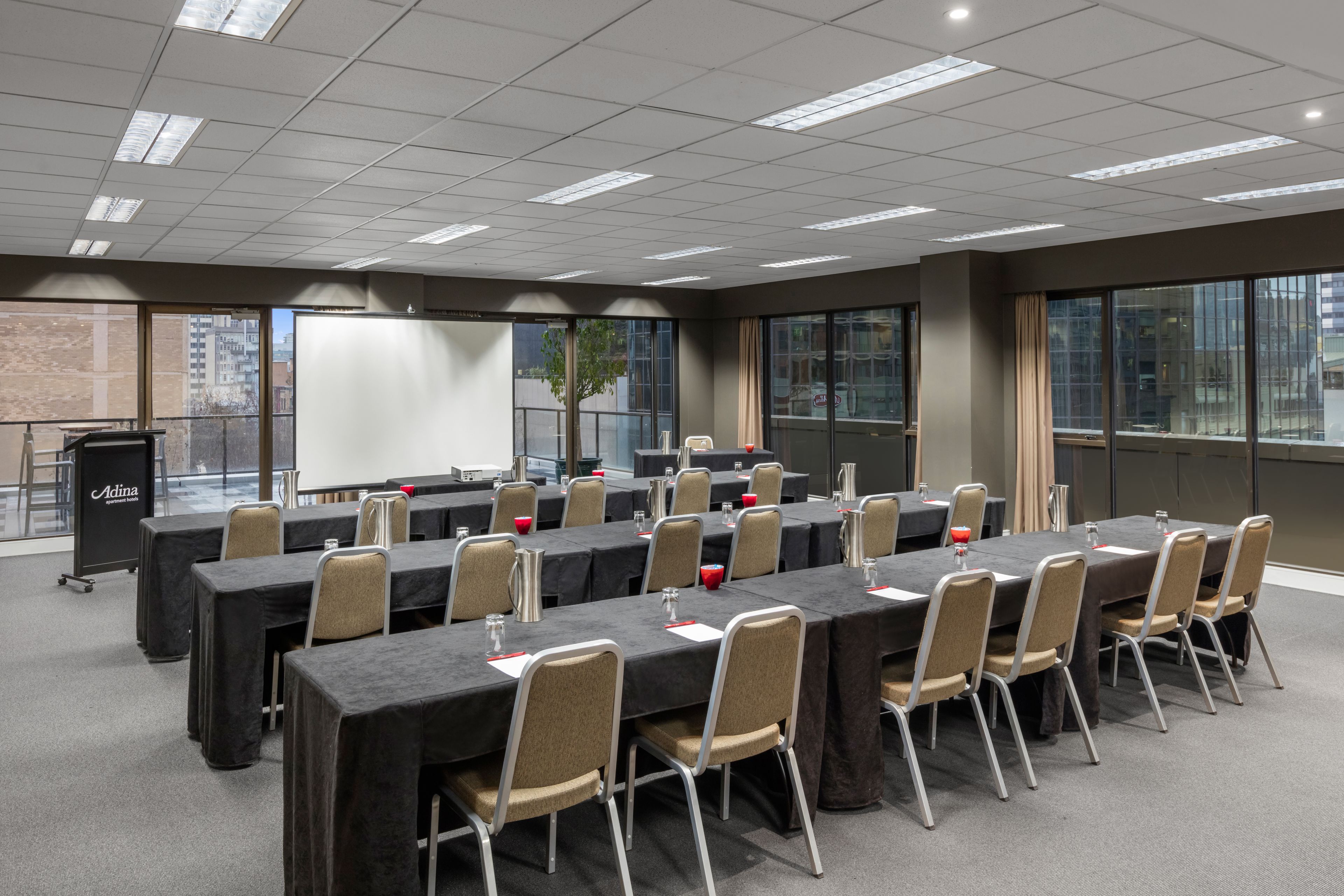 Une salle de conférence équipée de plusieurs tables et chaises, d’un écran de projection et d’une grande fenêtre donnant sur des immeubles.