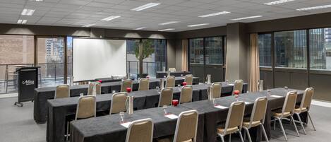 A conference room with multiple tables and chairs, a projector screen, and a large window with a view of buildings.