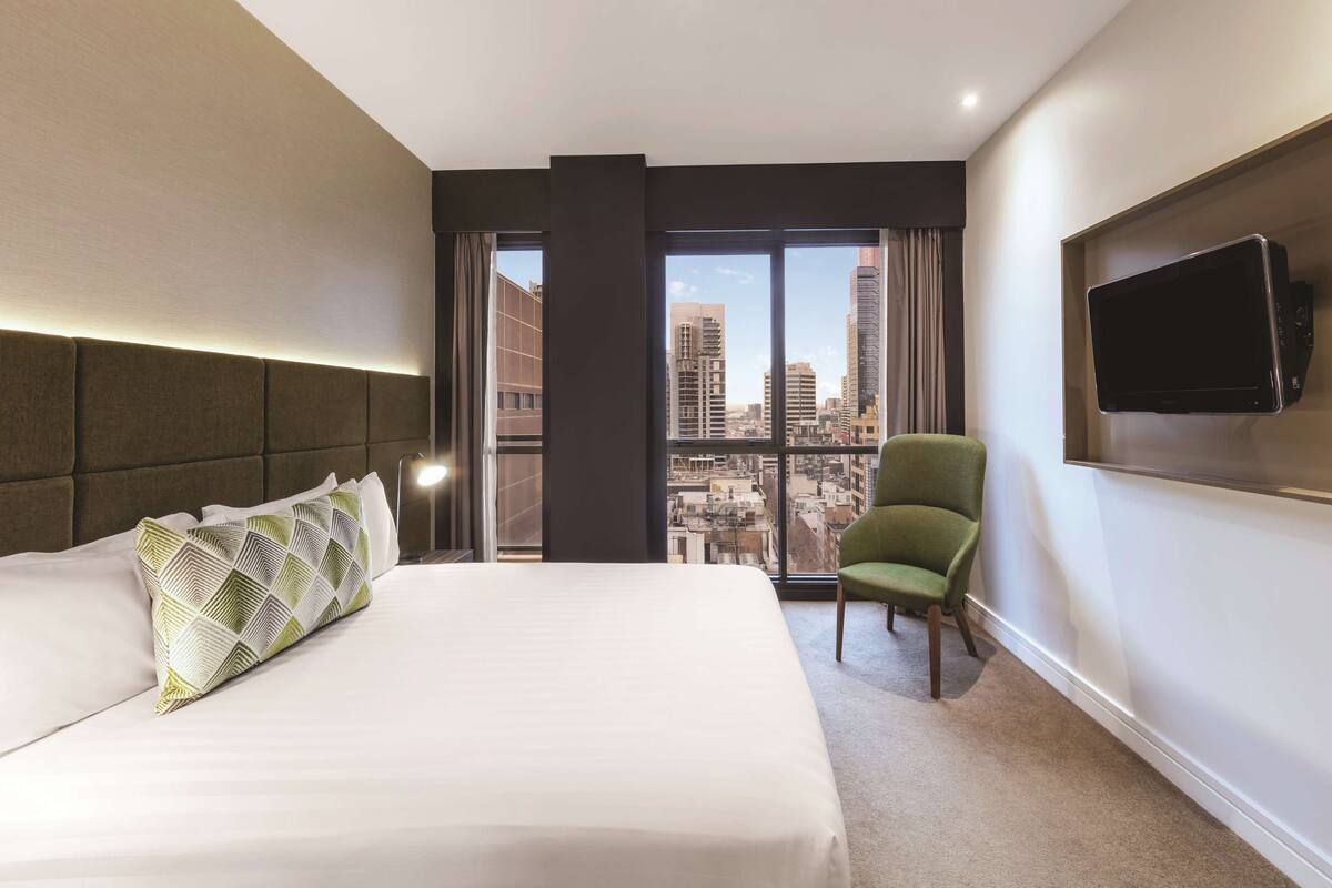 A hotel room with a large bed, a green armchair, a flat-screen TV mounted on the wall, and a city view through large windows.