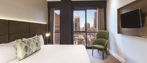 Une chambre d’hôtel avec un grand lit, un fauteuil vert, un téléviseur à écran plat fixé au mur et une vue sur la ville à travers de grandes fenêtres.
