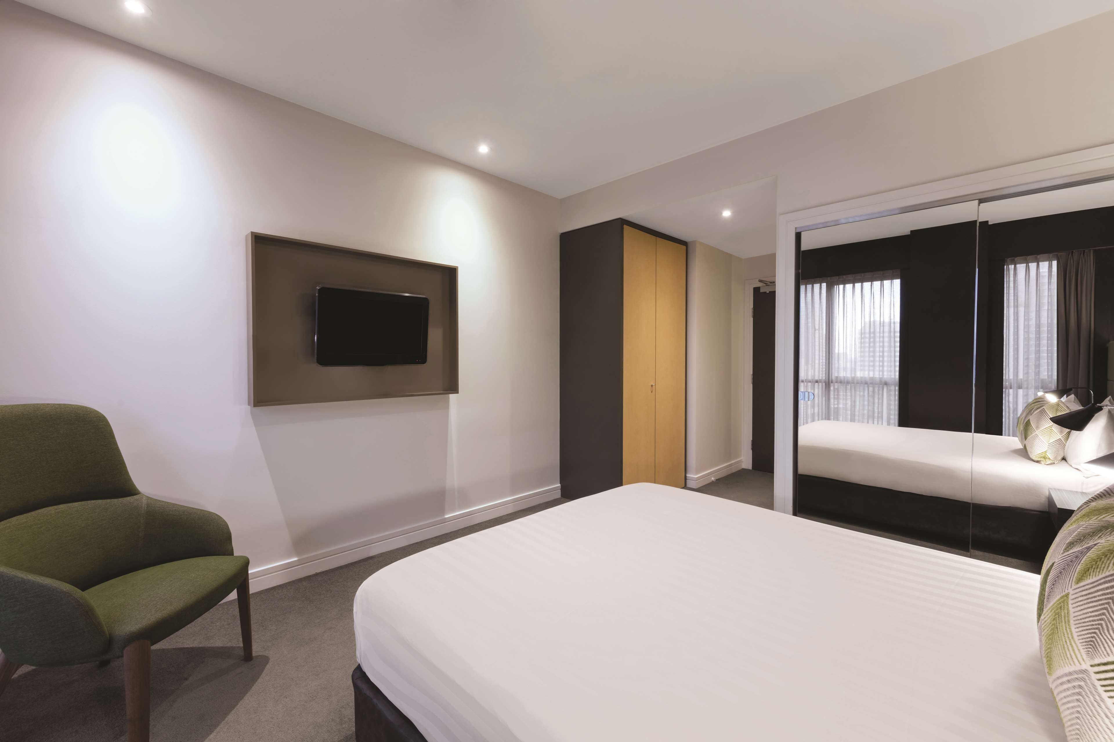 A modern hotel room with a bed, a green armchair, a TV mounted on the wall, and a sliding door leading to a balcony with a view.