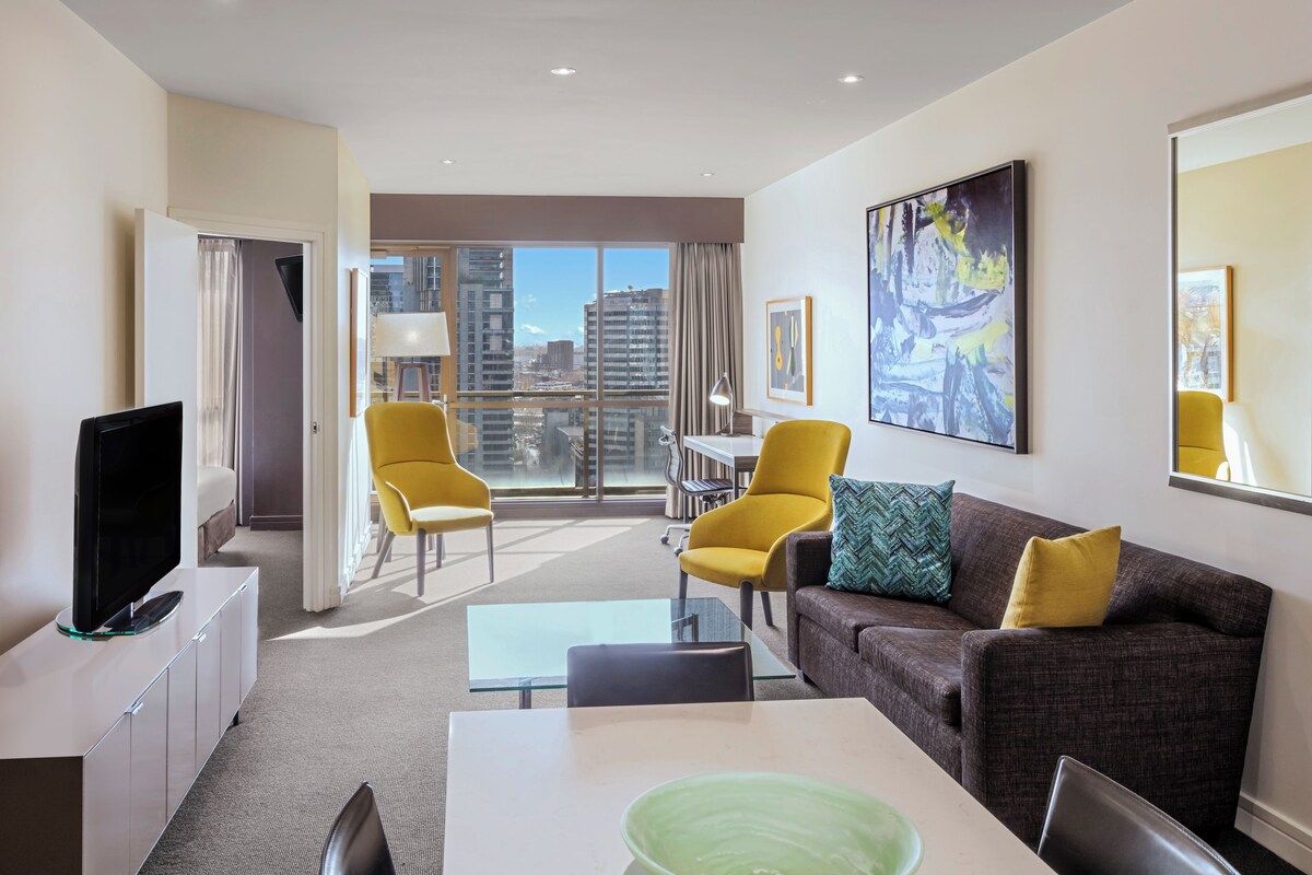 Una habitación de hotel moderna con televisión de pantalla plana, una mesa de comedor, un sofá y un amplio ventanal con vistas a la ciudad.