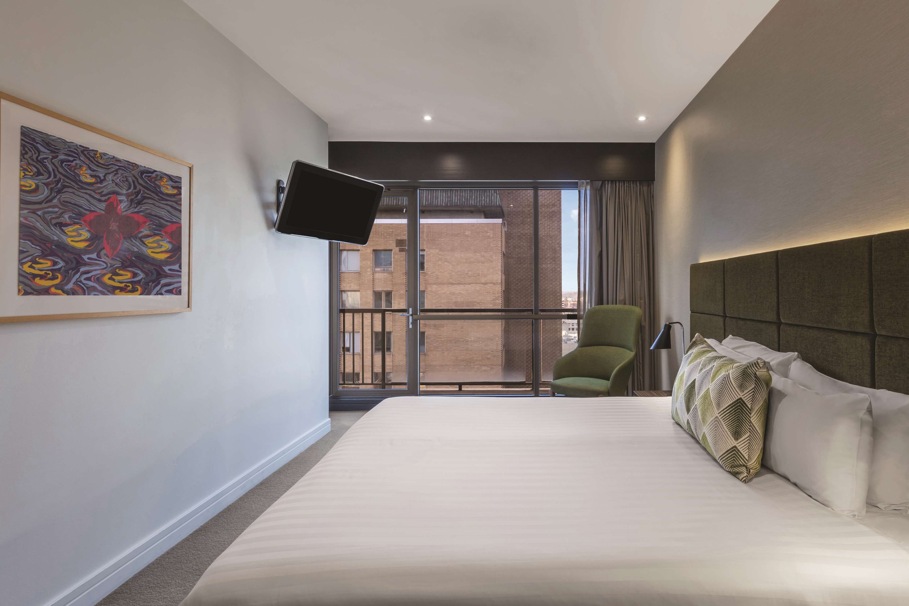 Habitación de hotel con una cama grande, un televisor montado, una silla verde y un balcón con vistas a edificios.