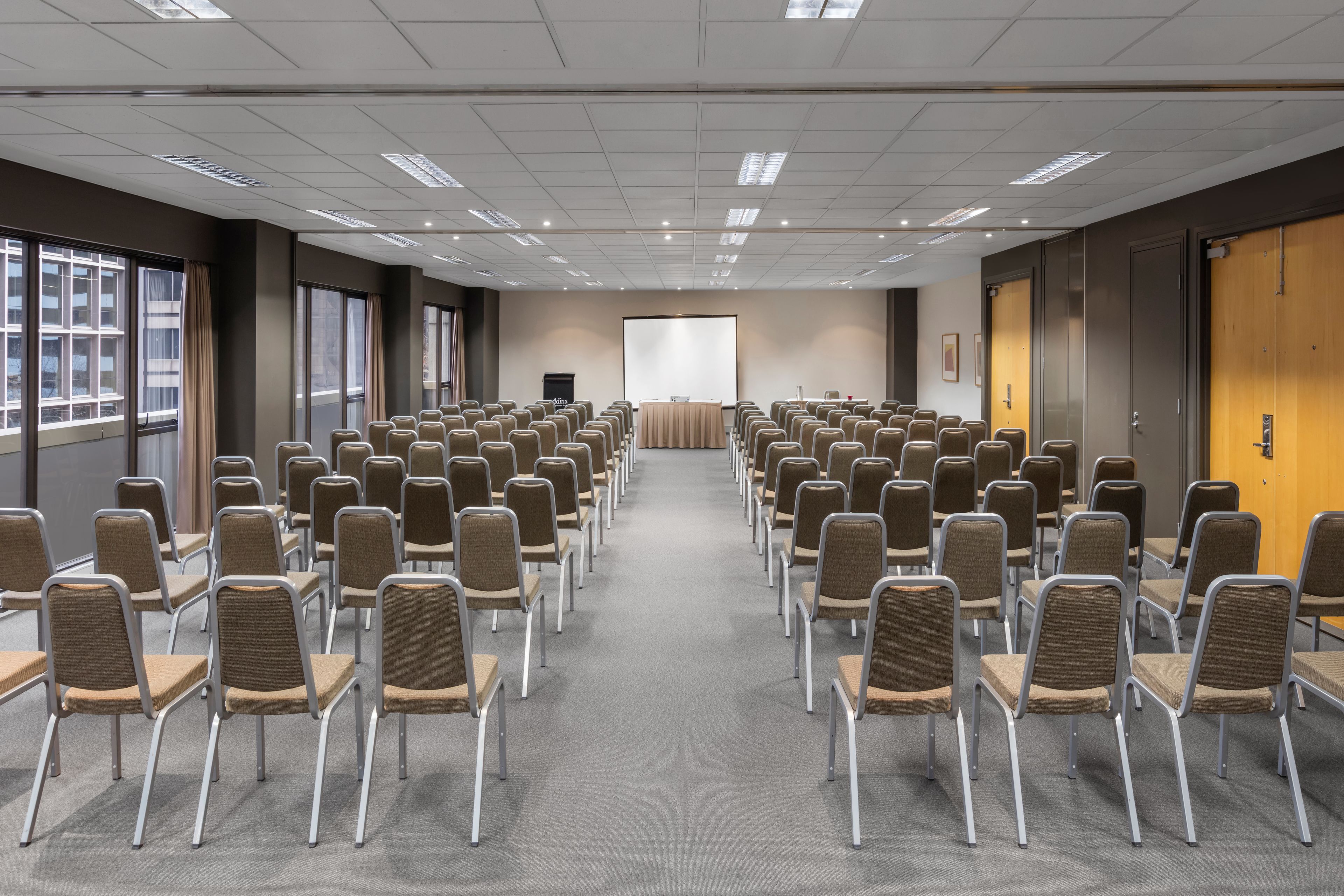 Une salle de conférence avec des rangées de chaises et de tables, un écran de projection et une grande fenêtre sur un des murs.