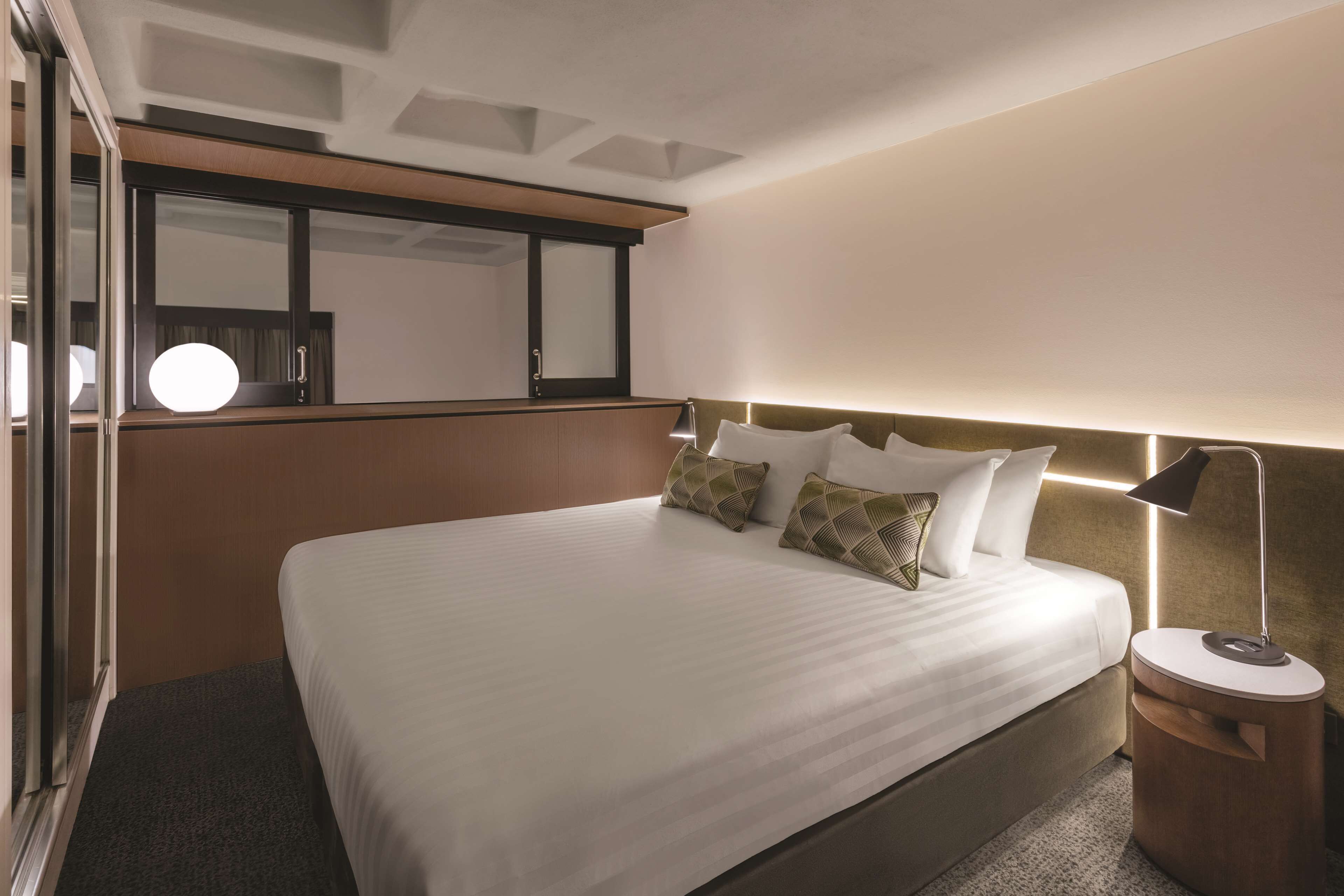 A hotel room with a large bed, a bedside table with a lamp, a mirror, and a window with a view of the sky.