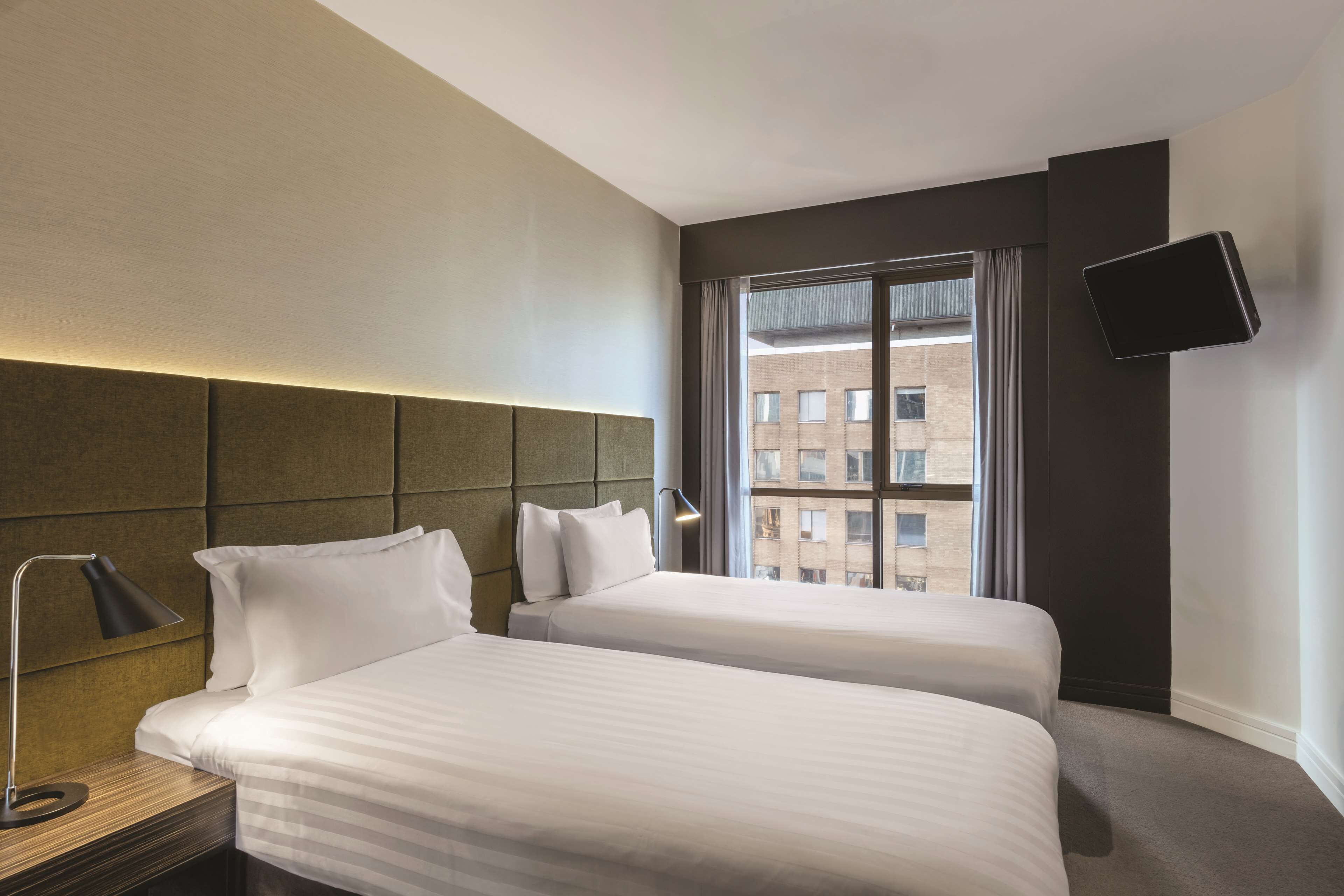 A hotel room with two beds, a bedside table with a lamp, a TV mounted on the wall, and a window with curtains.