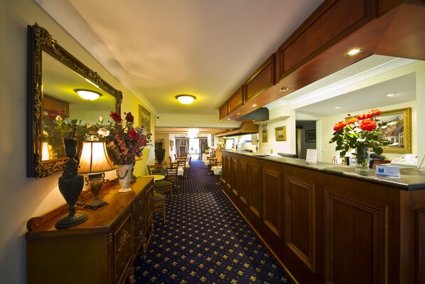 A hotel lobby with a patterned carpet, a reception desk, a large mirror, and a vase with flowers.