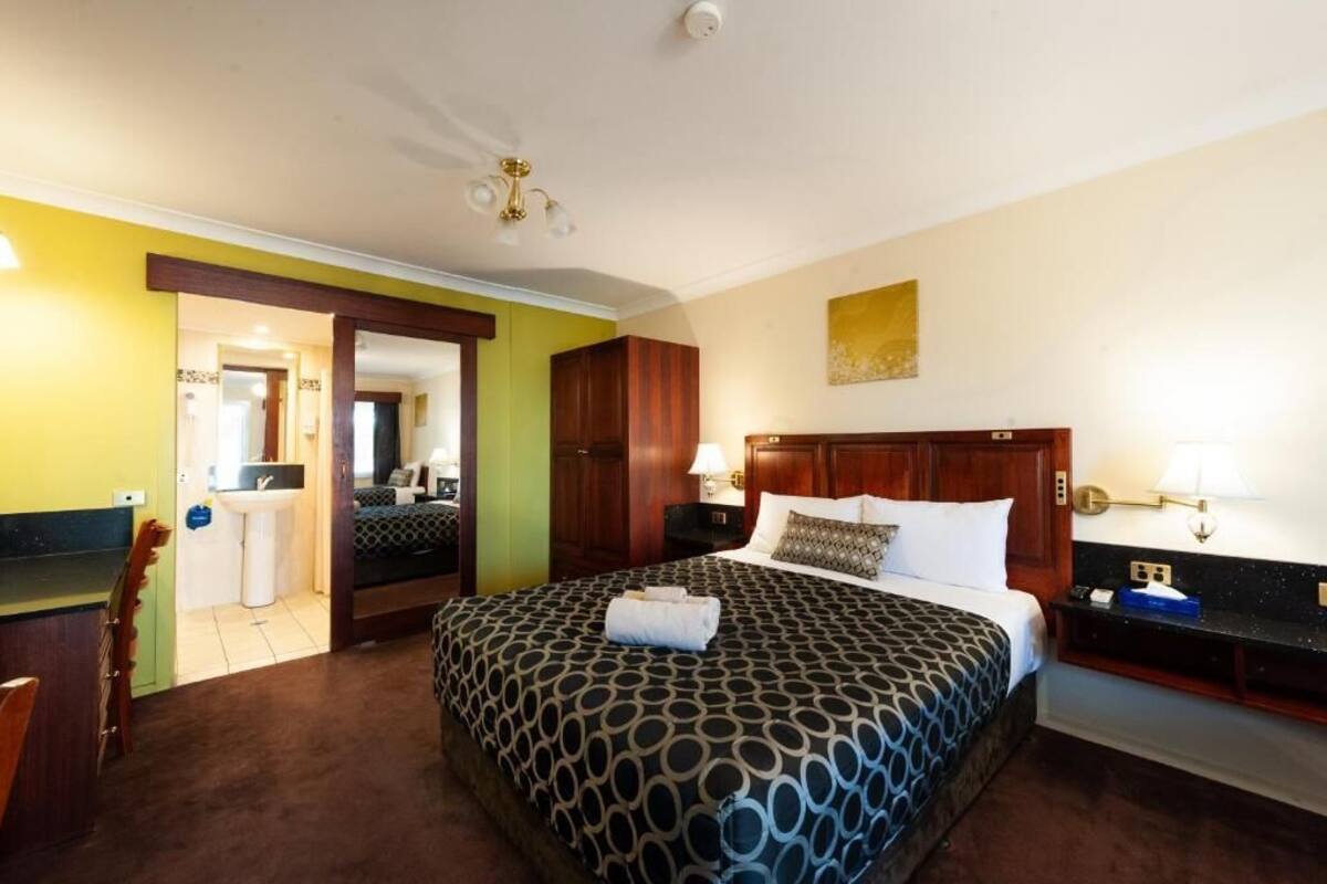 A hotel room with a large bed, a bedside table, a nightstand, a desk, a chair, a wardrobe, and a bathroom with a sink and mirror.