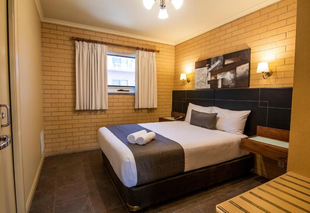 Hotelkamer met een bakstenen muur, een bed met hoofdeinde, twee nachtkastjes, een raam met gordijnen en een wandlamp.