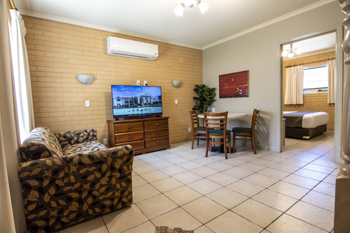 A living room with a sofa, a TV, a dining table with chairs, and a bed in the background.