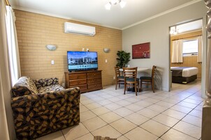 Una sala de estar con sofá, televisión, mesa de comedor con sillas y cama al fondo.