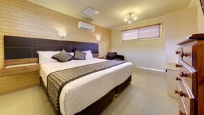 A hotel room with a large bed, a wooden headboard, a nightstand, a dresser, an air conditioning unit, and a window with blinds. - Albury Georgian Motel & Suites (Albury)
