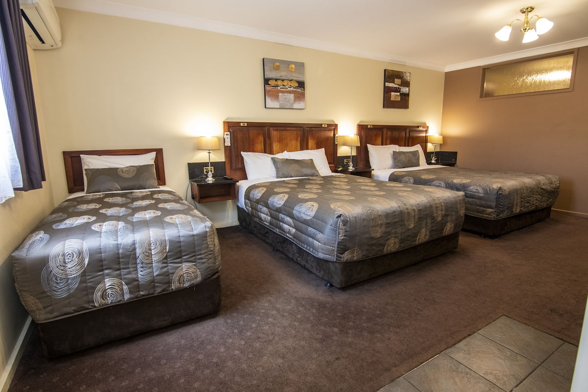 A hotel room with three beds, each with a patterned bedspread, a wooden headboard, and bedside lamps.